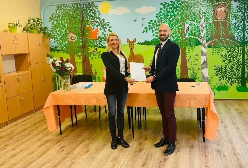 Zdjęcie wykonane w orientacji poziomej przedstawia kobietę i mężczyznę stojących wewnątrz sali przedszkolnej, którzy wspólnie trzymają biały dokument. Kobieta po lewej stronie ma na sobie czarną marynarkę, dżinsy oraz ciemne botki, a mężczyzna po prawej jest ubrany w ciemną marynarkę, bordowe spodnie i eleganckie obuwie. Przed nimi znajduje się stół przykryty pomarańczowym obrusem, na którym stoi wazon z bukietem kwiatów. Tło stanowi kolorowy mural przedstawiający leśny krajobraz z drzewami, sarną, wiewiórką i sową, co wskazuje na dziecięcą przestrzeń edukacyjną. Po lewej stronie kadru widoczne są wysokie, jasne szafki w zabudowie meblowej, które dopełniają wystrój pomieszczenia. Cała scena jest dobrze oświetlona i utrzymana w przyjaznej, formalnej atmosferze współpracy.