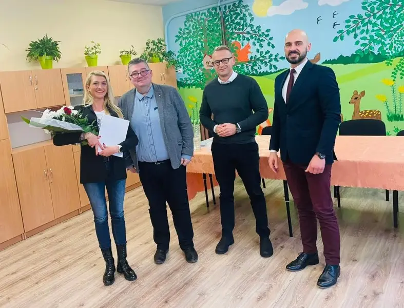 Zdjęcie przedstawia cztery uśmiechnięte osoby pozujące do fotografii wewnątrz pomieszczenia, prawdopodobnie w sali przedszkolnej lub szkolnej. Kobieta po lewej stronie trzyma bukiet kwiatów oraz dokument, a obok niej stoi trzech mężczyzn w strojach typu business casual oraz formalnych. Układ zdjęcia jest horyzontalny, a postacie znajdują się w centrum kadru na tle jasnych szafek oraz ściany ozdobionej kolorowym malowidłem z motywami przyrodniczymi i zwierzętami. W tle widoczny jest również stół nakryty brzoskwiniowym obrusem, co sugeruje przygotowanie do spotkania lub uroczystości. Jasna kolorystyka wnętrza, drewniana podłoga i przyjazna atmosfera budują profesjonalny, lecz ciepły charakter fotografii, która może pełnić funkcję relacji z wydarzenia społecznego lub zawodowego.