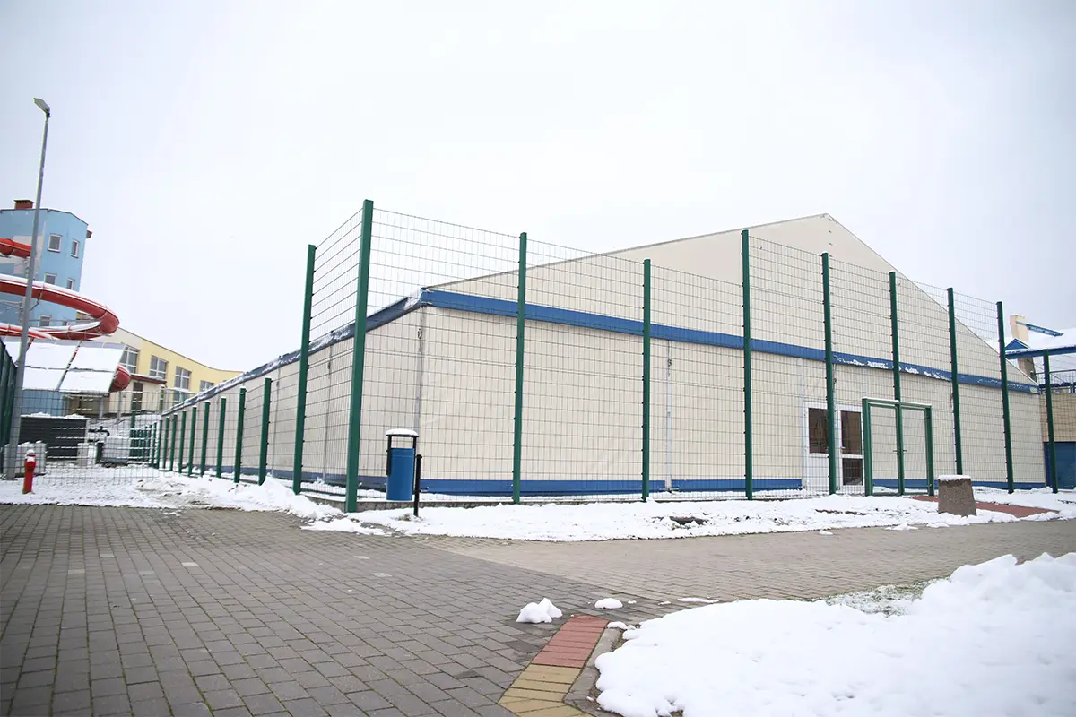 Zdjęcie przedstawia dużą halę namiotową z jasnym poszyciem, która jest otoczona wysokim ogrodzeniem z zielonej siatki panelowej. Obiekt znajduje się na otwartym terenie pokrytym częściowo śniegiem, w otoczeniu wyłożonym kostką brukową, co nadaje mu charakteru zimowej infrastruktury sportowo-rekreacyjnej. W lewym górnym rogu widać fragment kolorowej zjeżdżalni wodnej oraz niebieski budynek, co sugeruje sąsiedztwo aquaparku lub centrum sportowego. Układ kadru jest panoramiczny, z wyraźnym podziałem na pierwszy plan z ośnieżoną nawierzchnią i dalszy plan z bryłą namiotu, co sprawia wrażenie szerokiej perspektywy zewnętrznej. Niebo jest pochmurne, co tworzy stonowane, zimowe oświetlenie podkreślające teksturę materiału hali oraz stalową konstrukcję ogrodzenia.